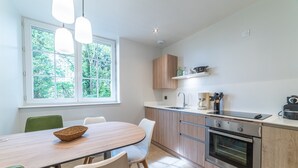 A modern kitchen with a wooden table, white chairs, a built-in oven, and a window with a view of greenery. - Le Jungle - Appt pour 6 avec parking privé (Paray-le-Monial)