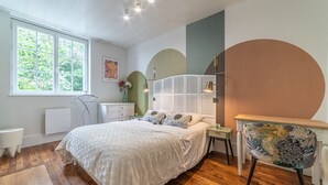 A bedroom with a large bed, a wooden floor, a window with a view of trees, a white radiator, a bedside table, and a chair with a patterned backrest. - Le Jungle - Appt pour 6 avec parking privé (Paray-le-Monial)