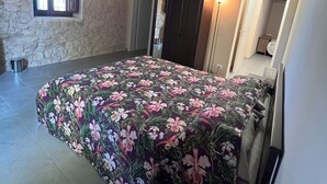 A bedroom with a floral bedspread, a stone wall, a window with shutters, a door, and a ceiling light.
