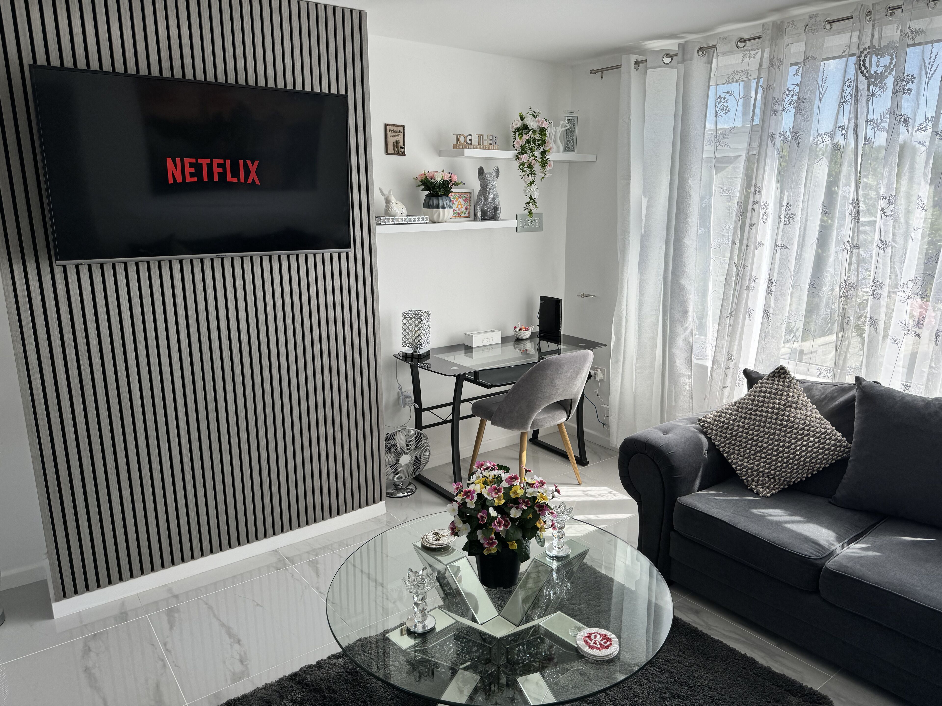 A modern living room with a flat-screen TV displaying 'NETFLIX', a glass coffee table with decorative items, a grey sofa, and a dining area with a glass table and chairs.