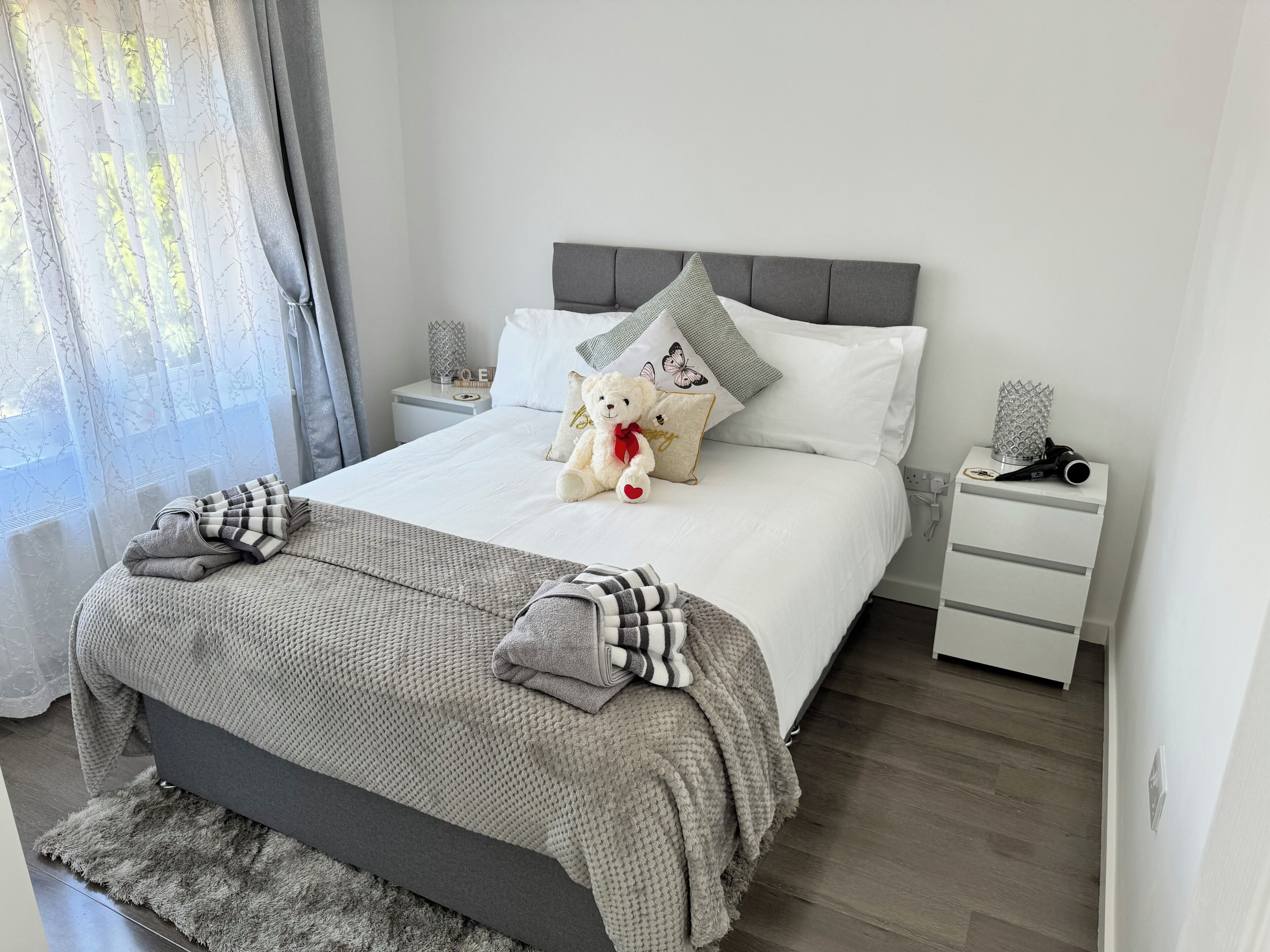 A neatly made bed with a grey blanket and pillows, a bedside table with a lamp, and a window with sheer curtains.