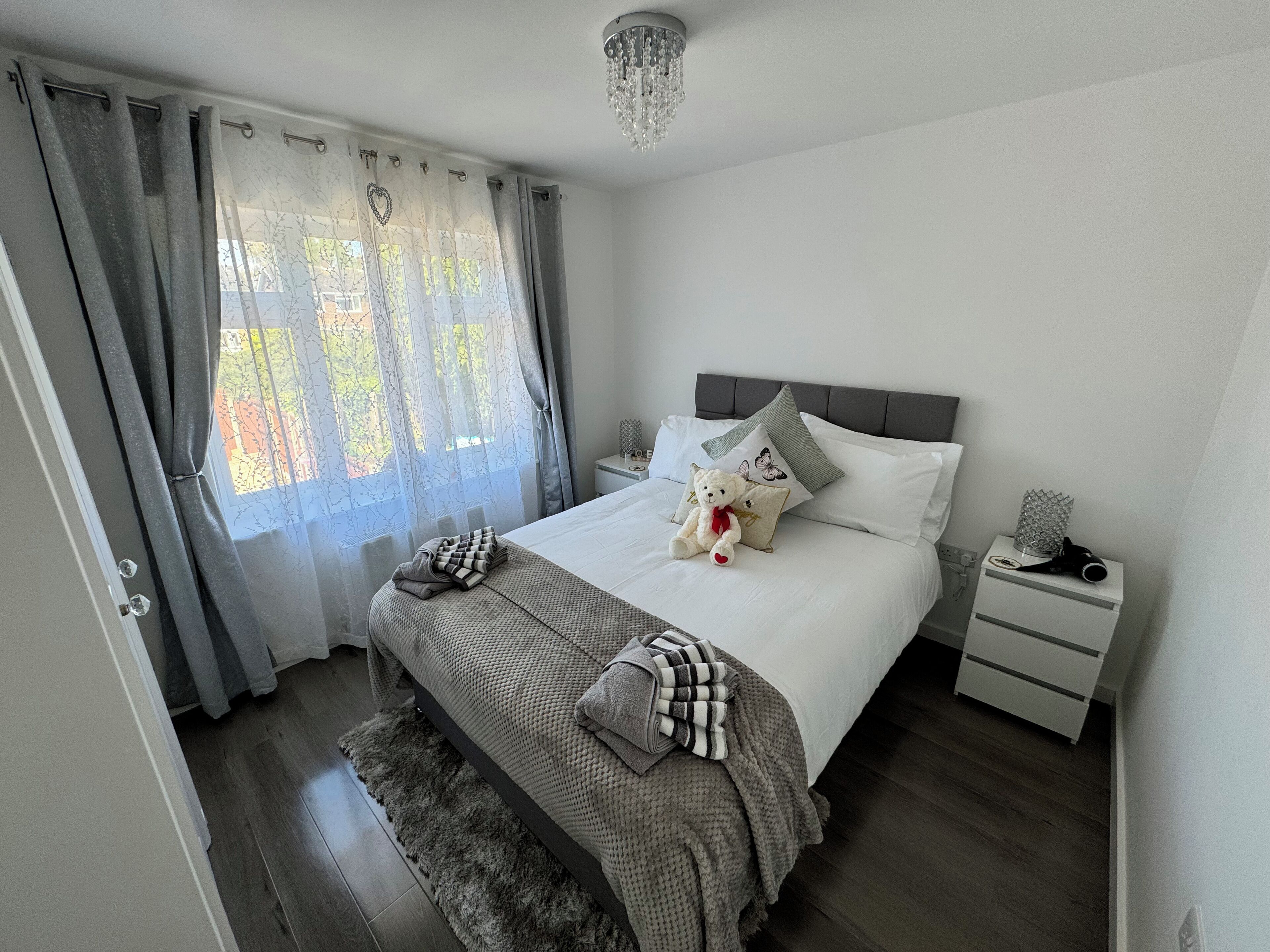 A neatly made bed with a teddy bear, a grey blanket, and a heart-shaped pillow. There is a nightstand with a lamp and a small decorative item.