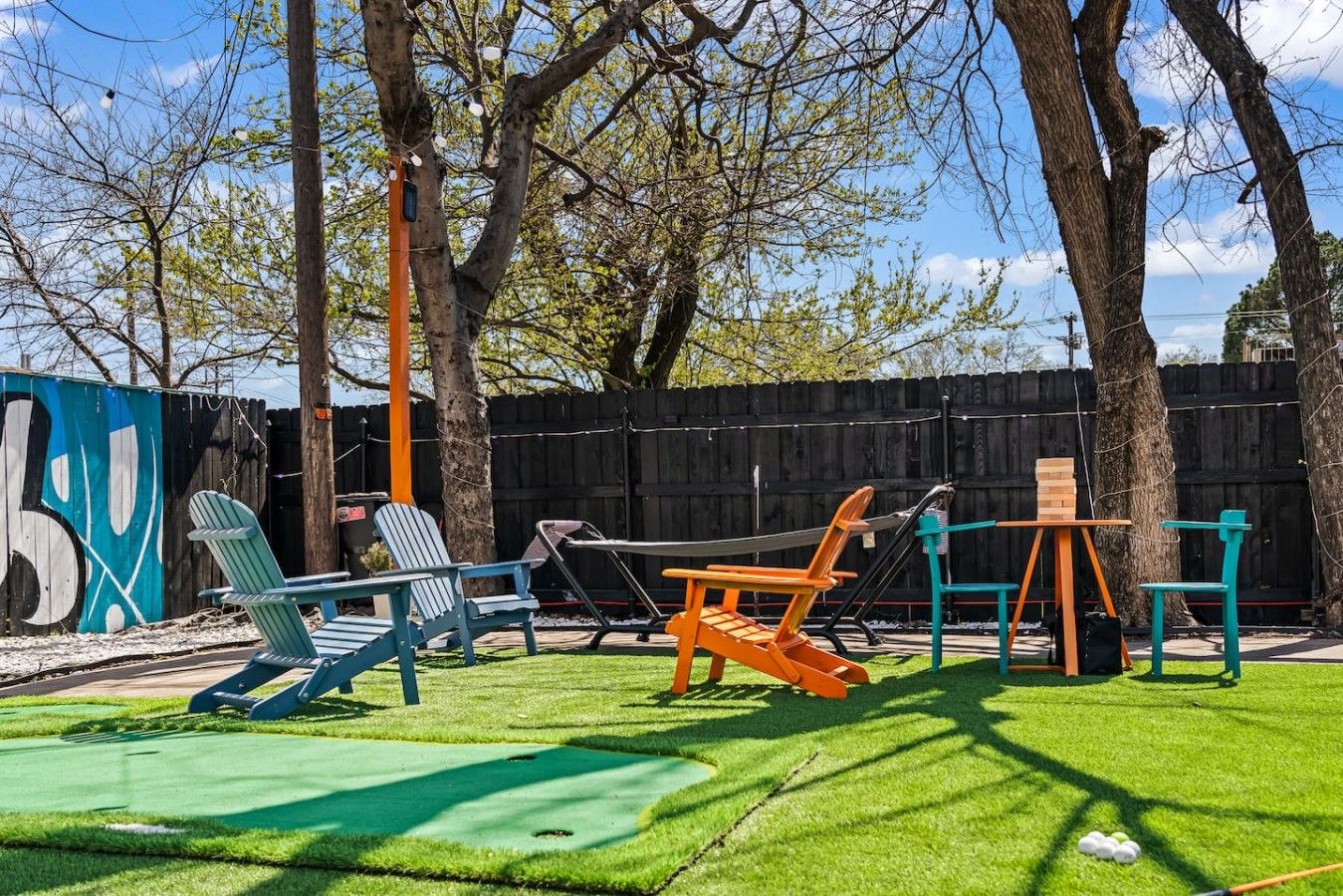An outdoor seating area with Adirondack chairs and a table, surrounded by a wooden fence and trees.