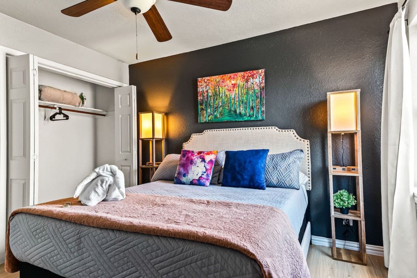 A bedroom with a bed, a ceiling fan, a painting, bedside lamps, and a nightstand.