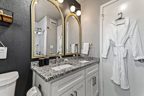 A bathroom with a double sink, a large mirror with lights, a towel rack, and a robe hanging on the wall.
