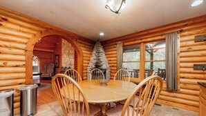 A wooden cabin interior with a round dining table surrounded by wooden chairs, a stone fireplace, and a window with curtains. - Family & Dog Friendly Escape w/ 2 Hot Tubs (McHenry)