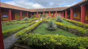 A courtyard with a central fountain, surrounded by neatly trimmed hedges and plants, and a tiled roof structure with red pillars.