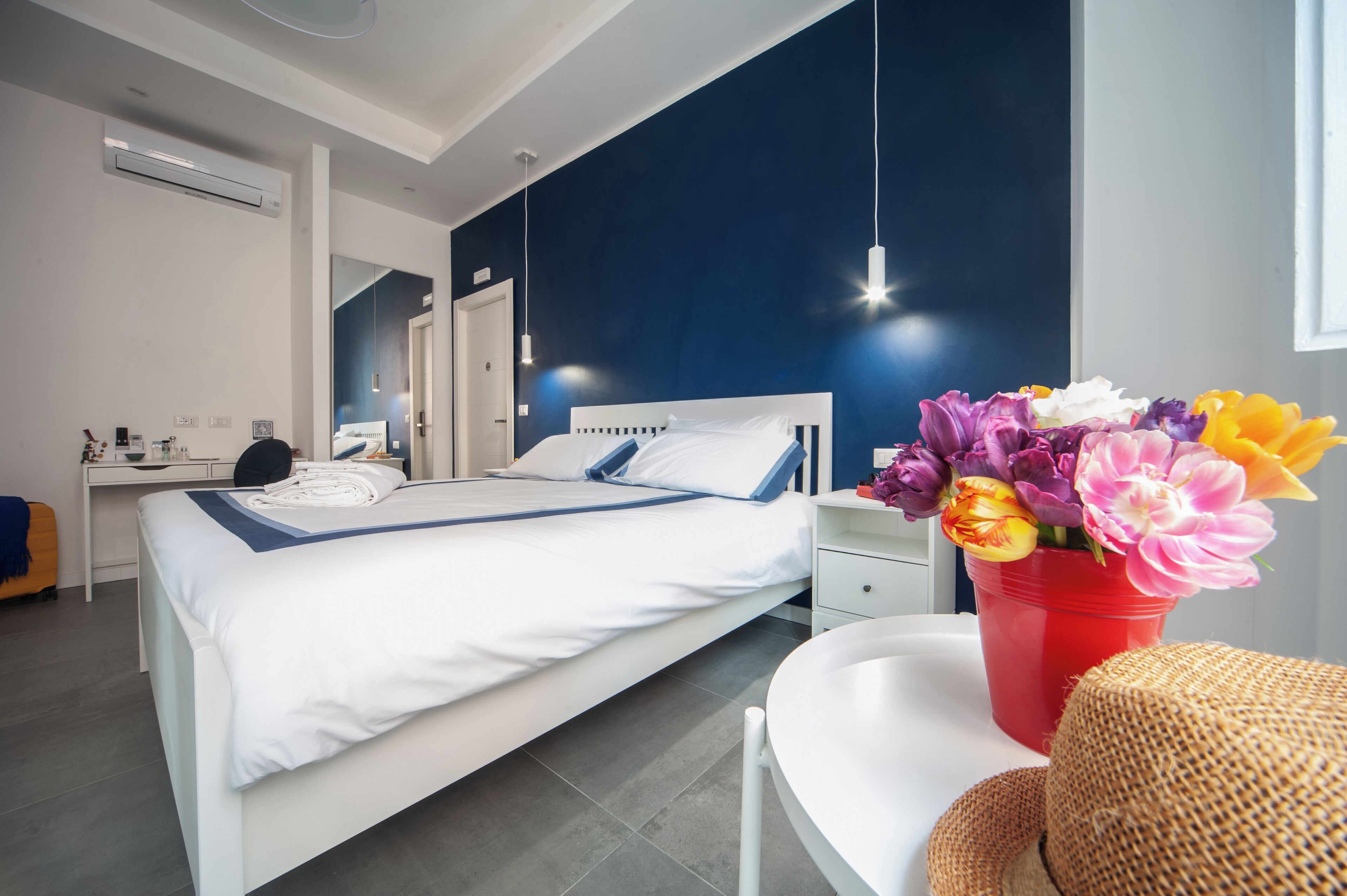 Quarto de hotel moderno com uma cama bem arrumada, uma escrivaninha com cadeira, um vaso de flores vermelho e um espelho grande.