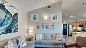 Una sala de estar moderna con sofá, sillón y mesa de centro, además de una zona de cocina con electrodomésticos y obras de arte en las paredes.