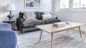 A modern living room with a grey sofa, a small side table with a lamp, a coffee table with books, and a large window with a view of buildings. - Impeccable 2-bed Apartment in London (London)