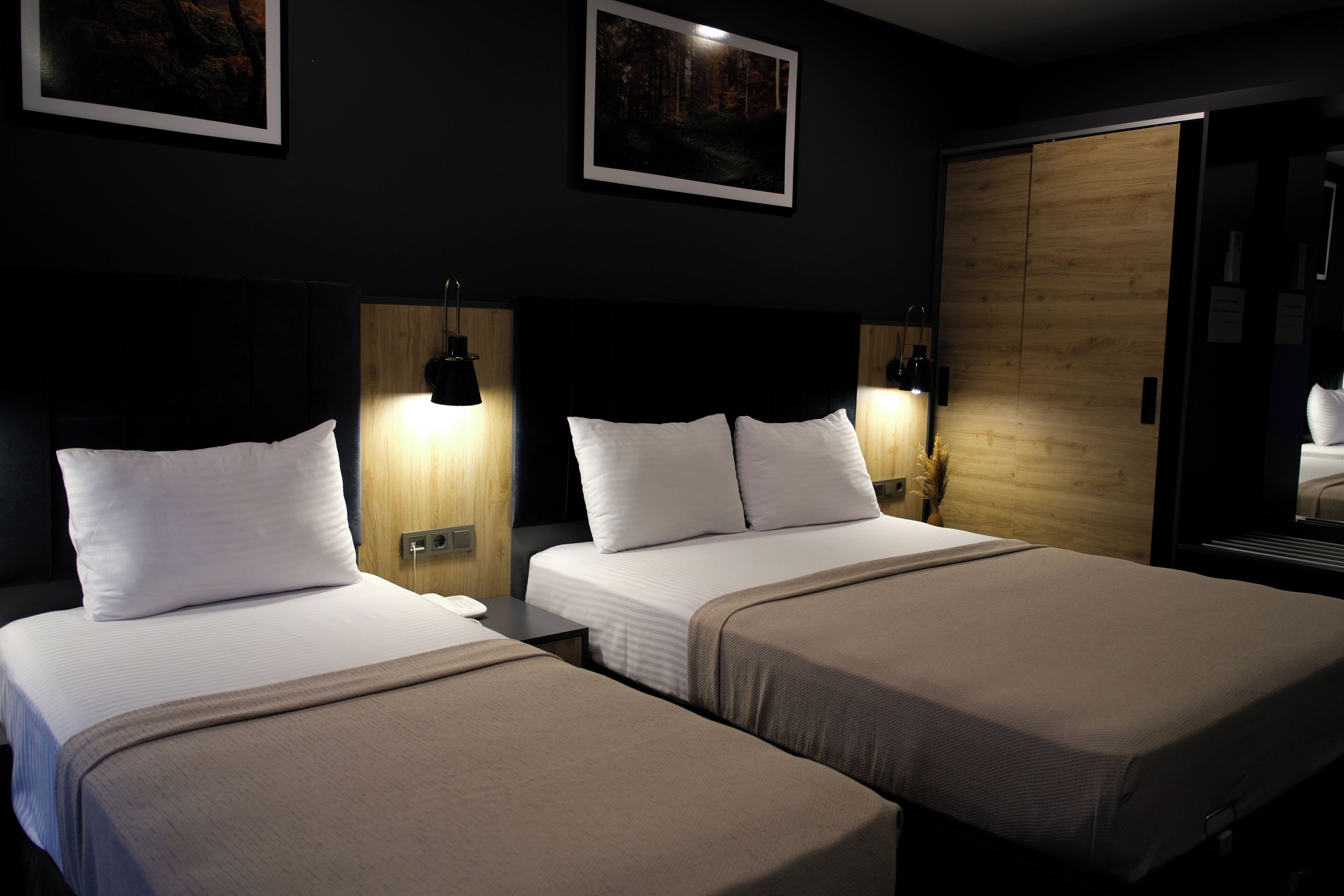 A hotel room with two beds, each with white pillows and brown bedspreads. There are wall-mounted lamps on either side of the beds, and a framed picture above the beds.