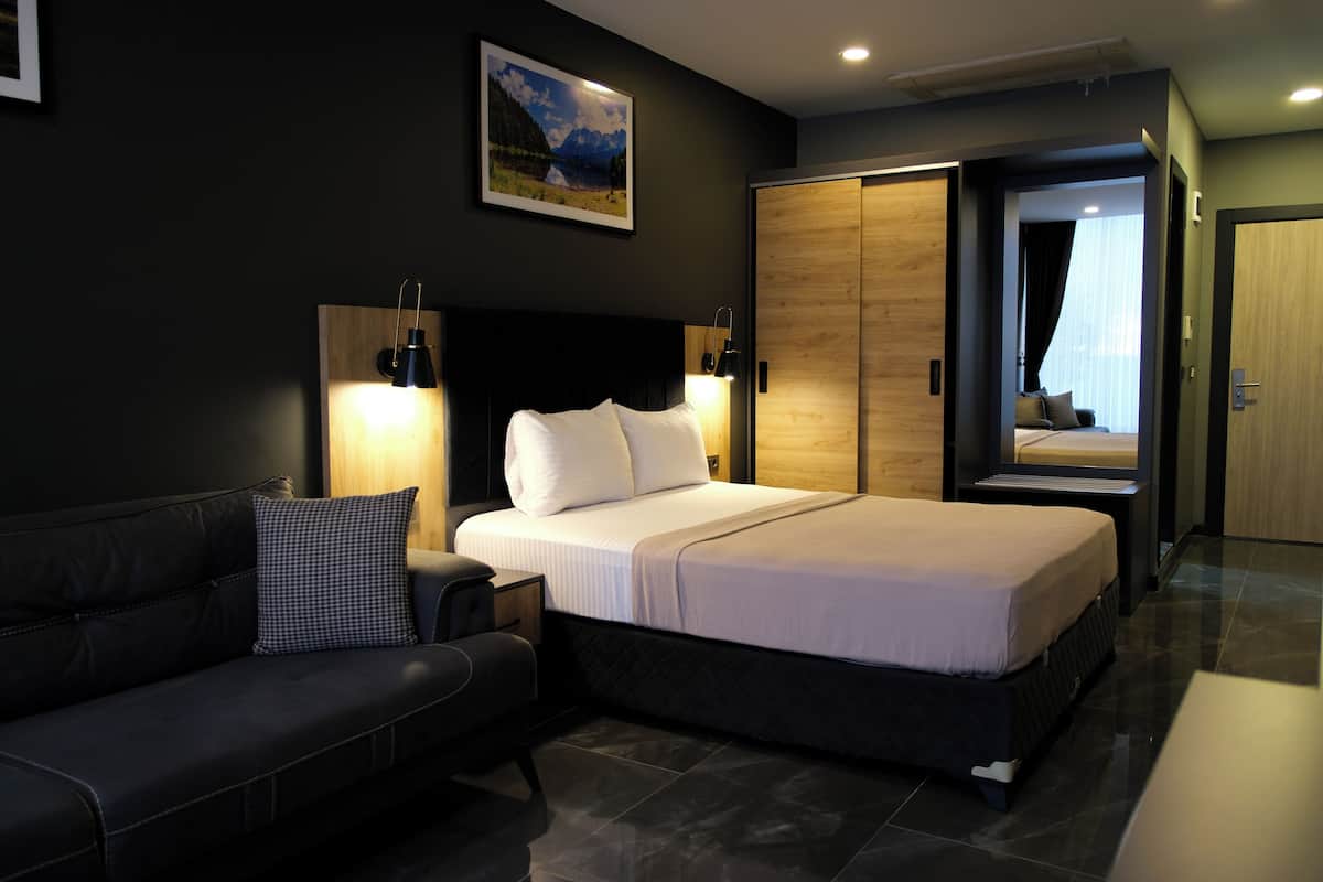A modern hotel room with a large bed, a dark-colored sofa, a wooden wardrobe, and a framed picture on the wall.