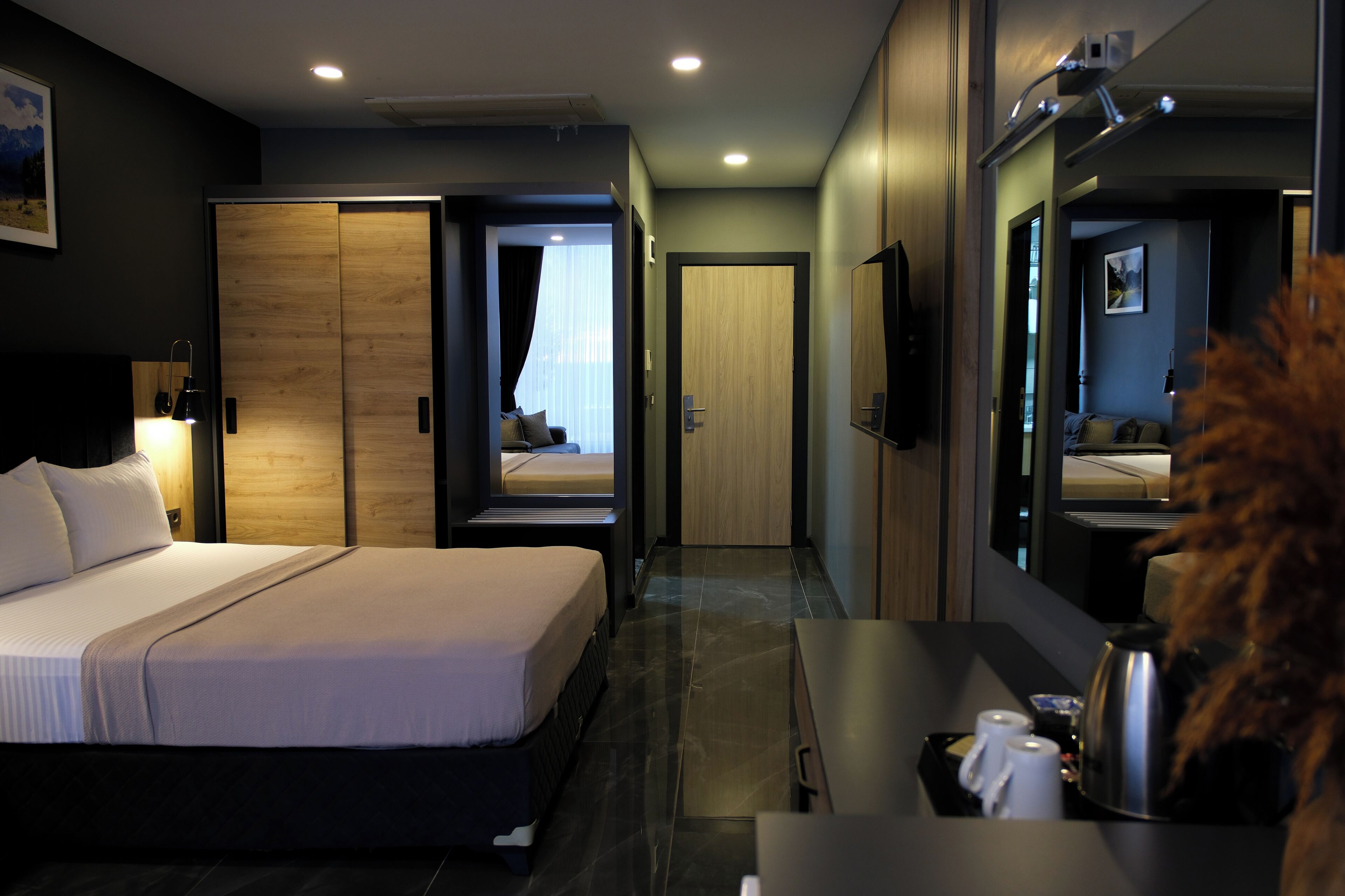 A modern hotel room with a large bed, a desk with a kettle, and a mirror.