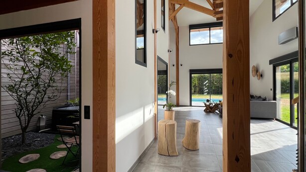 A modern interior with a wooden beam structure, a glass door leading to a pool area, and a seating arrangement with a view of the outdoors.