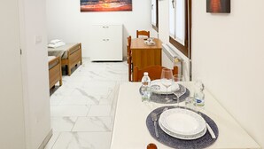 A compact hotel room with a dining area, a bed, a small cabinet, and a painting of a sunset.
