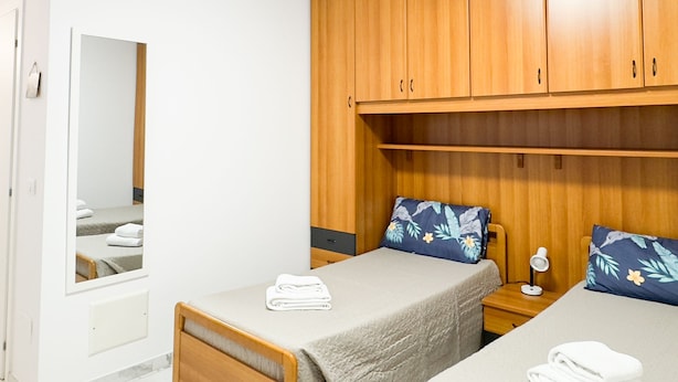 A bedroom with two single beds, wooden wardrobes, a bedside table with a lamp, and a mirror on the wall.