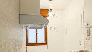 A compact kitchen with white tiles, a built-in stove, a hanging light fixture, and a window with wooden shutters.