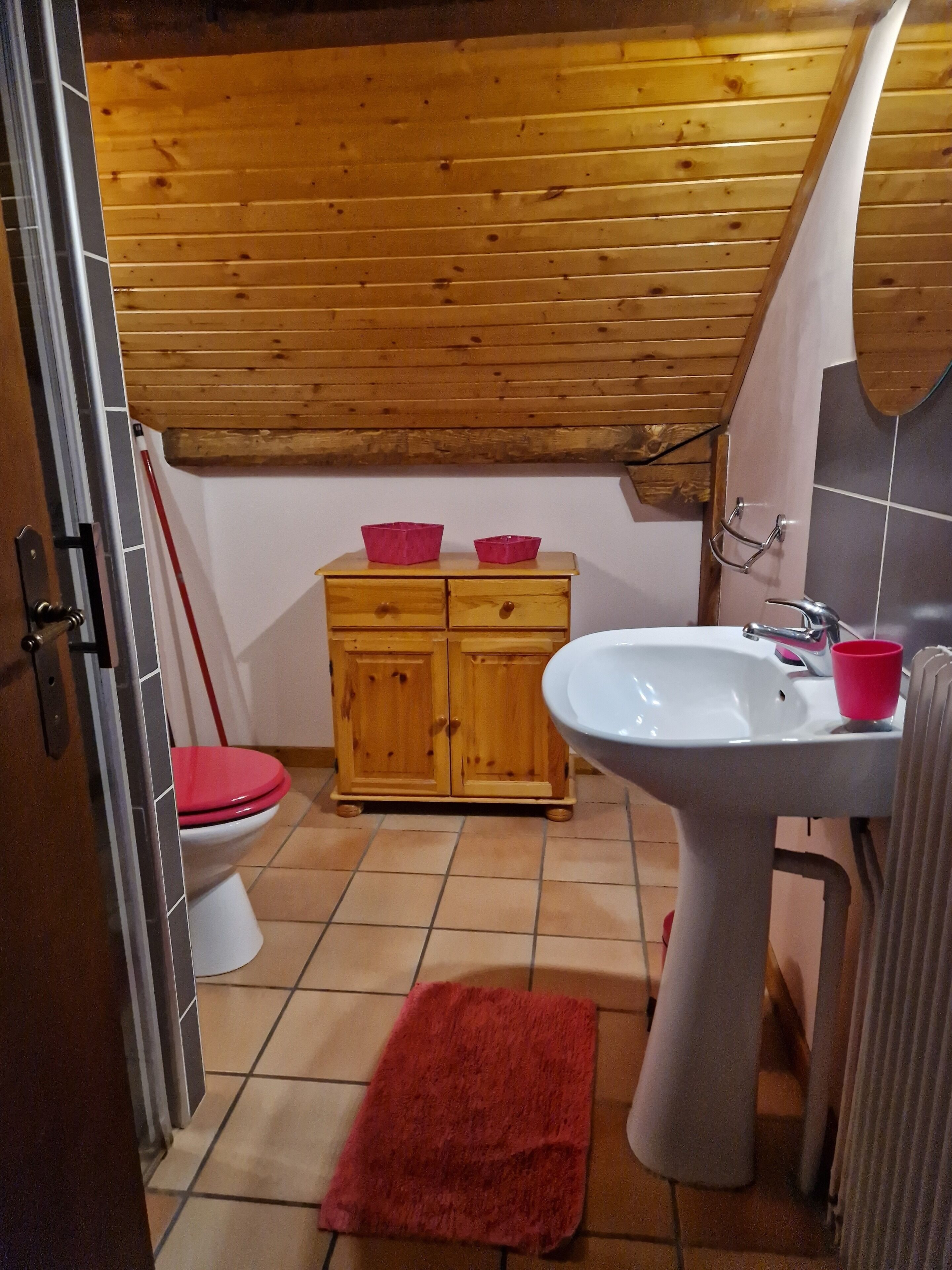 Pieni kylpyhuone, jonka seinät on verhoiltu puupaneeleilla. Siellä on wc, puinen kaappi, jossa on kaksi vaaleanpunaista säilytysastiaa, valkoinen pesuallas ja punainen pyyhe lattialla.