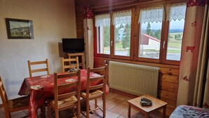 Une pièce confortable avec une table et des chaises en bois, un téléviseur, un radiateur et une fenêtre avec des rideaux.