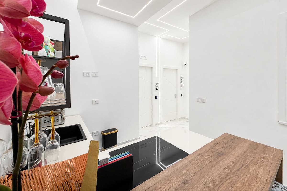 Quarto de hotel moderno com mesa de jantar de madeira, tapete preto, parede branca e uma orquídea rosa.