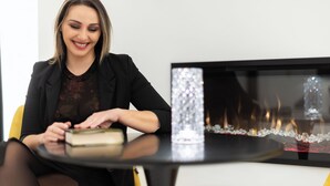 Una mujer sentada a una mesa con un libro, un jarrón decorativo y una chimenea al fondo.