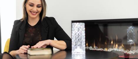 A woman sitting at a table with a book, a decorative vase, and a fireplace in the background.