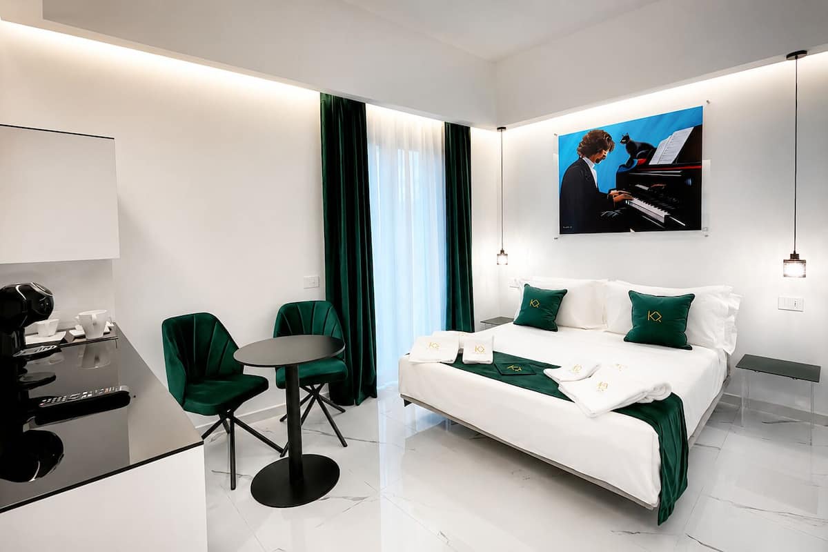 Quarto de hotel moderno com cama, escrivaninha com cafeteira, duas cadeiras e um quadro de um homem tocando piano.