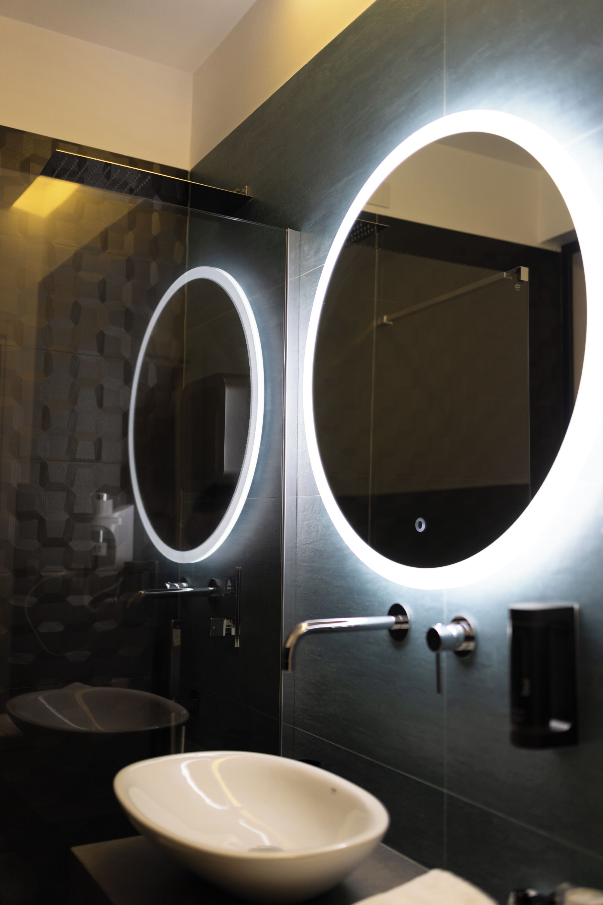 Un baño moderno con un espejo ovalado grande, un lavabo blanco y una pared revestida con baldosas oscuras.