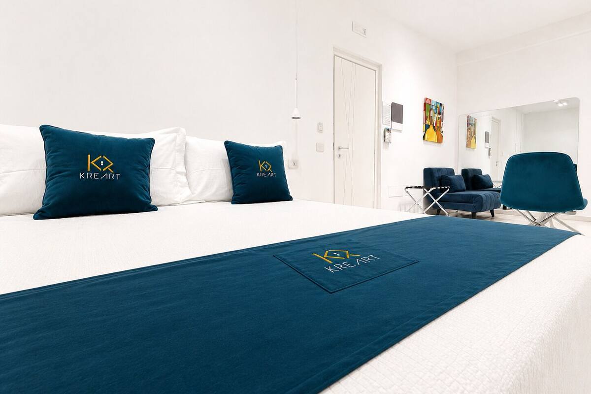 Quarto de hotel com uma cama grande, duas almofadas com logotipo, um cobertor azul, uma cadeira e uma escrivaninha.