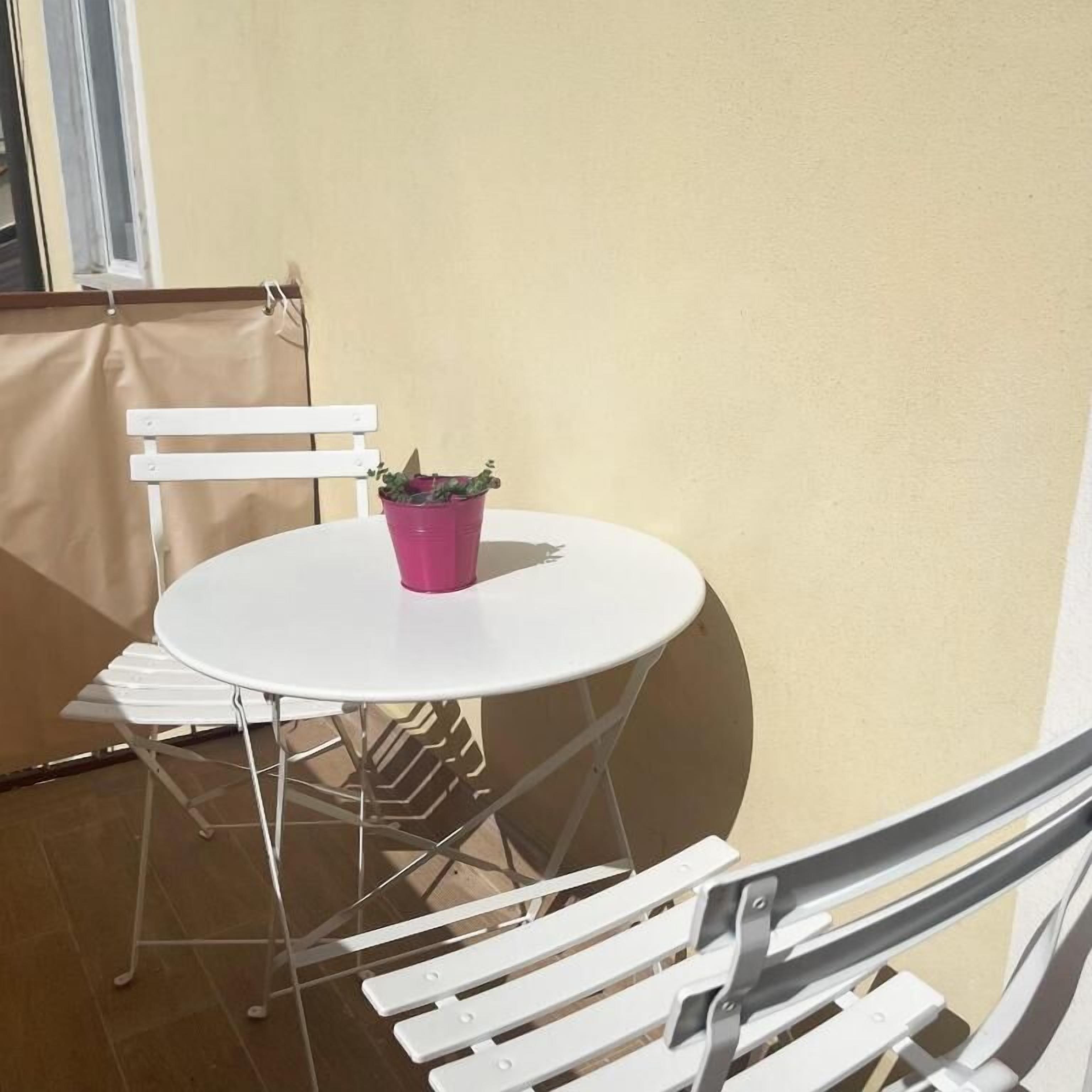 A small outdoor seating area with a round table and white chairs, a potted plant on the table, and a white chair leaning against a wall.