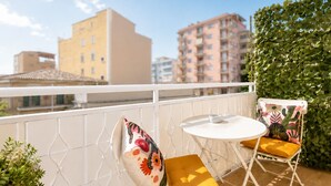 Un balcón con barandilla blanca, una mesita redonda pequeña, dos sillas con cojines y vistas a edificios al fondo.