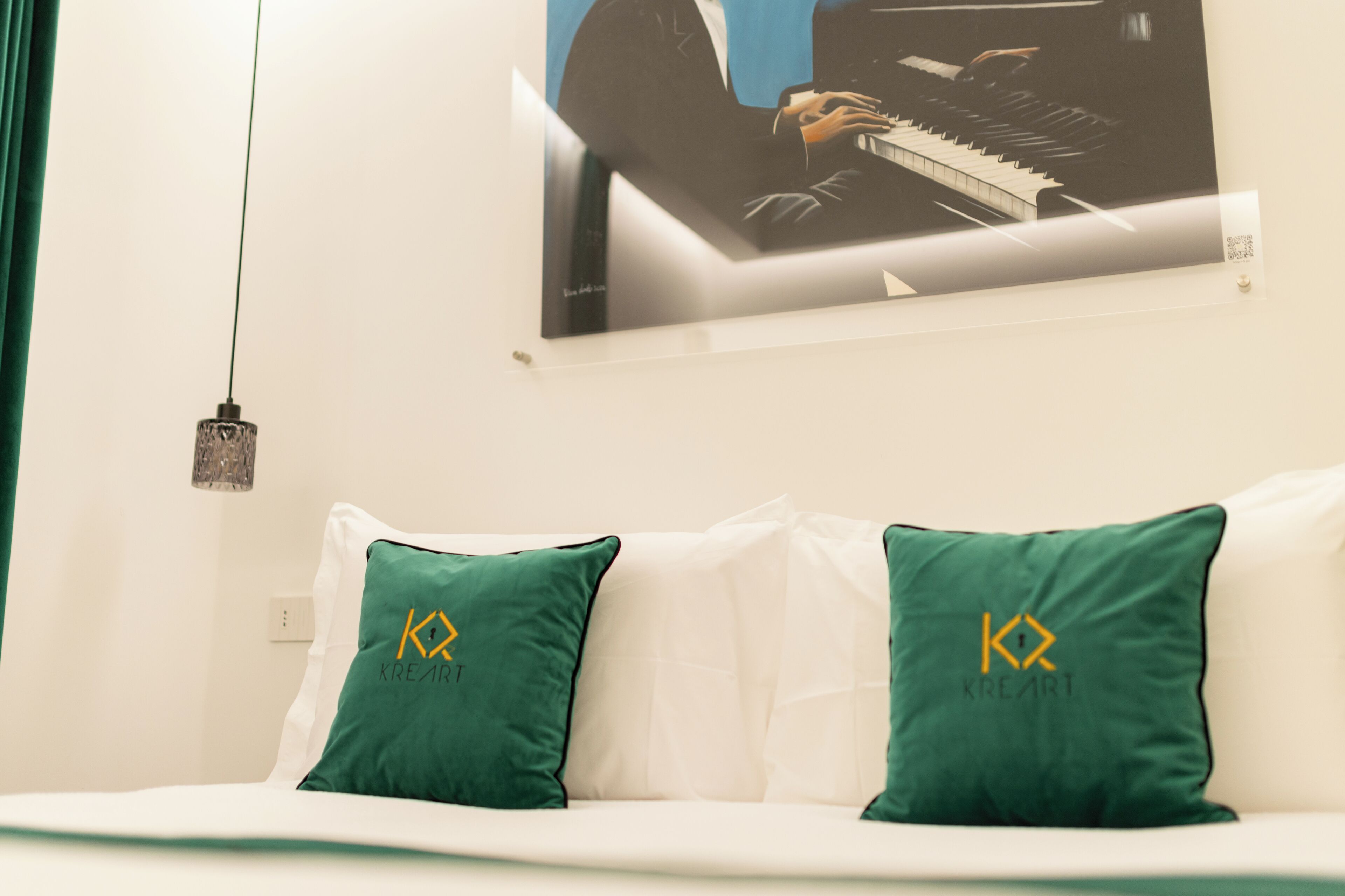 Een hotelkamer met een witte bank, twee groene kussens met een logo, een schilderij van een persoon die piano speelt, en een hanglamp.