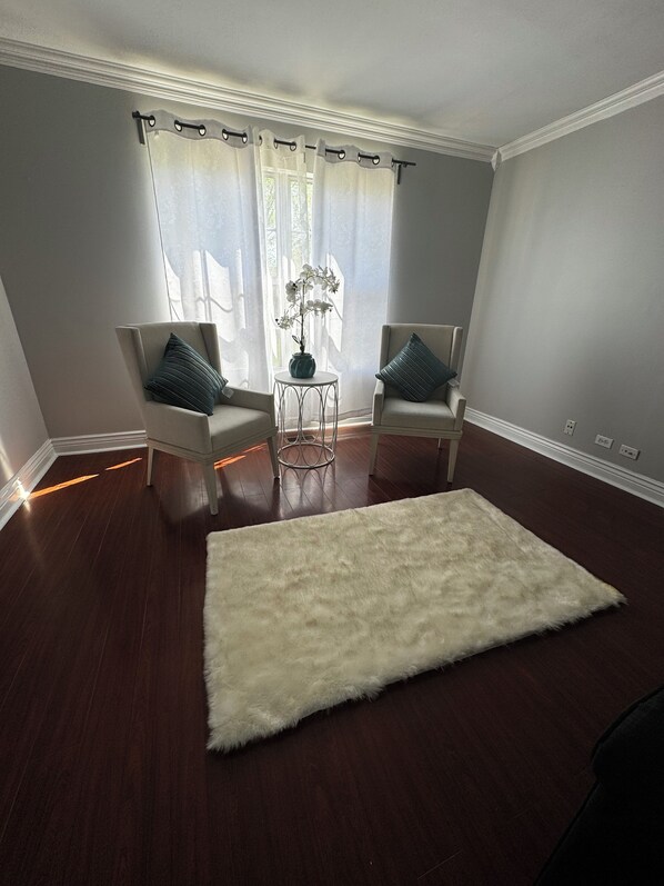 Una sala de estar con dos sillones, un tapete blanco mullido, una mesita redonda con un florero y una ventana con cortinas de encaje.