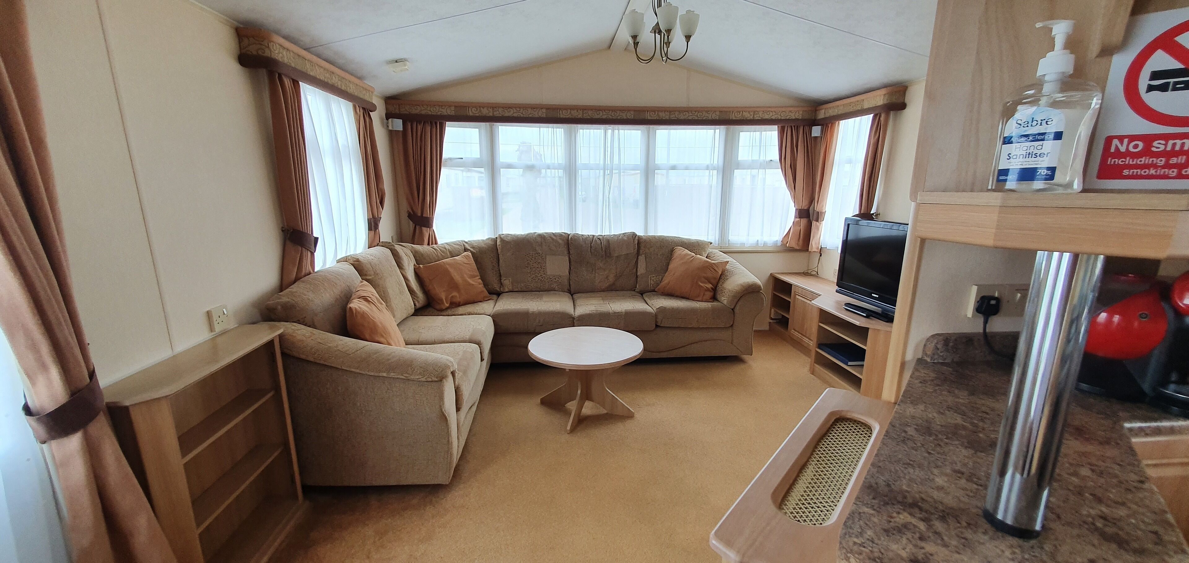 A cozy living room with a sofa, a small round table, and a television. There is a shelf with a hand sanitizer bottle and a sign indicating no smoking.