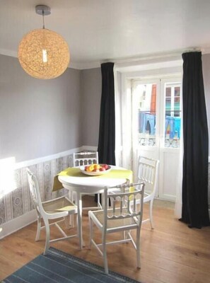 En matplats med ett runt bord, vita stolar, en hängande lampa och utsikt genom ett fönster med gardiner.