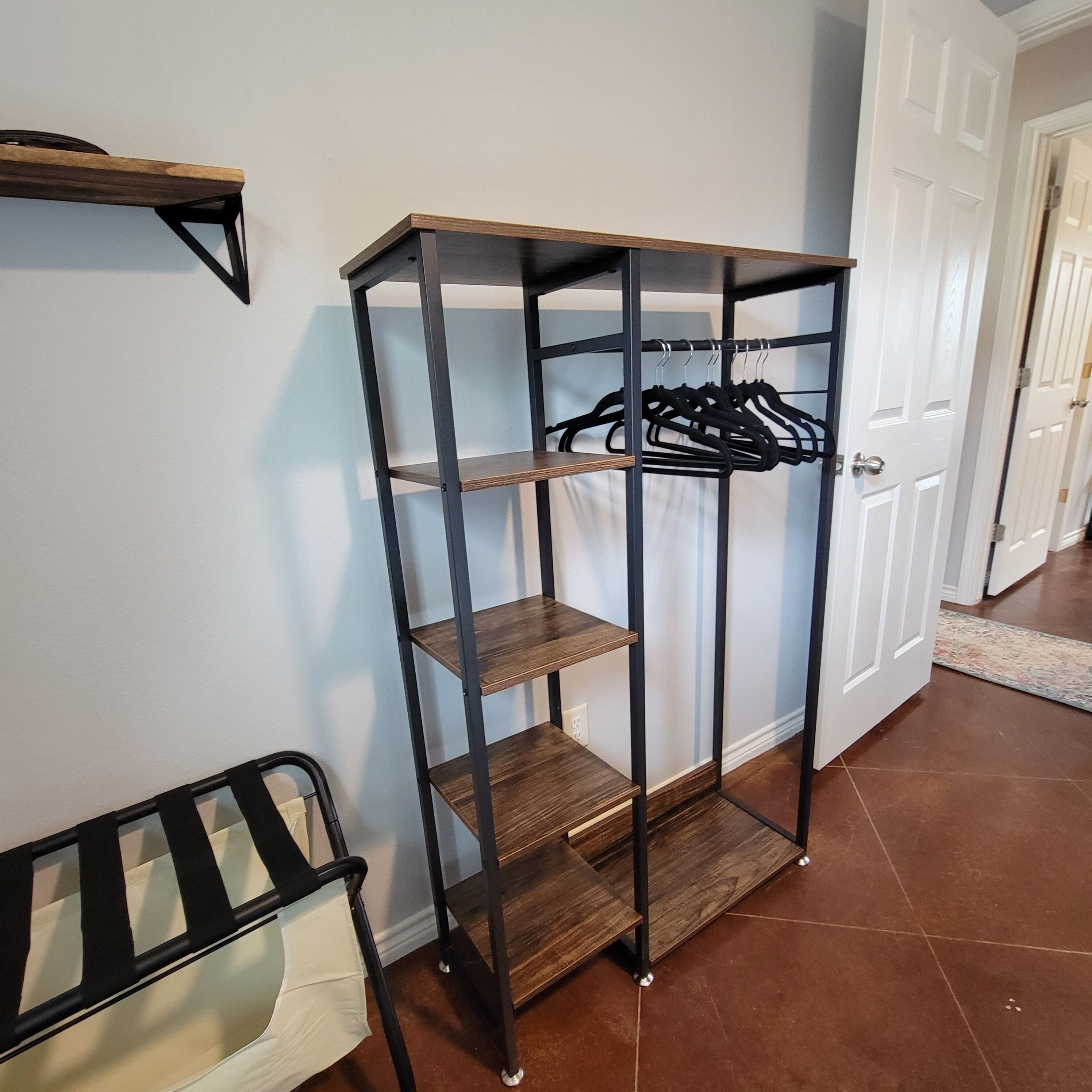 A black metal shelf unit with three shelves and a hanging rack, positioned against a white wall with a door in the background.