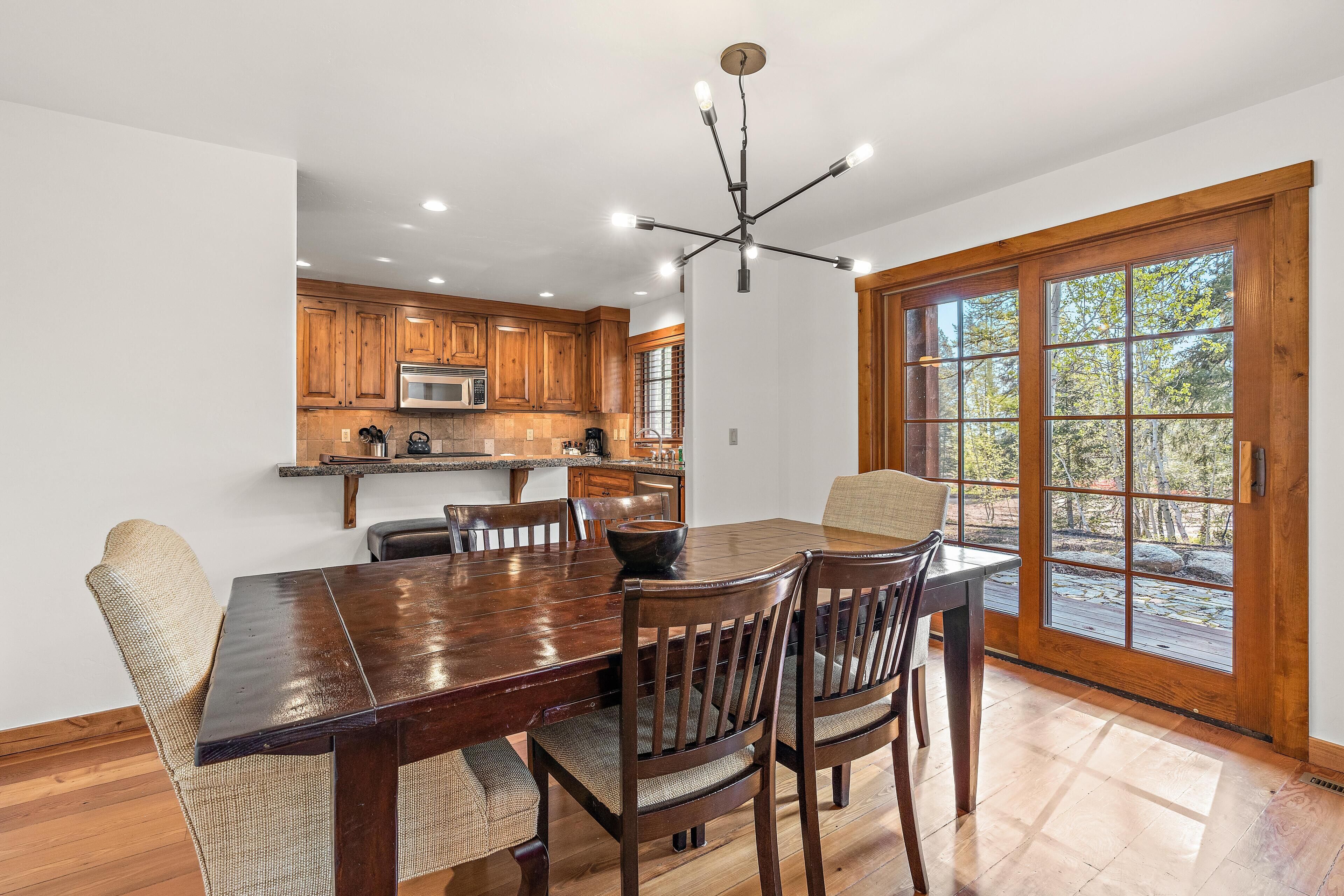A dining area with a wooden table and chairs, a kitchen in the background, and a large window with a view of trees.