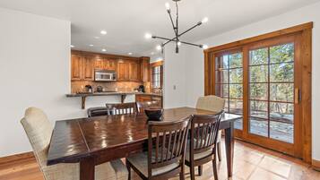 A dining area with a wooden table and chairs, a kitchen in the background, and a large window with a view of trees.