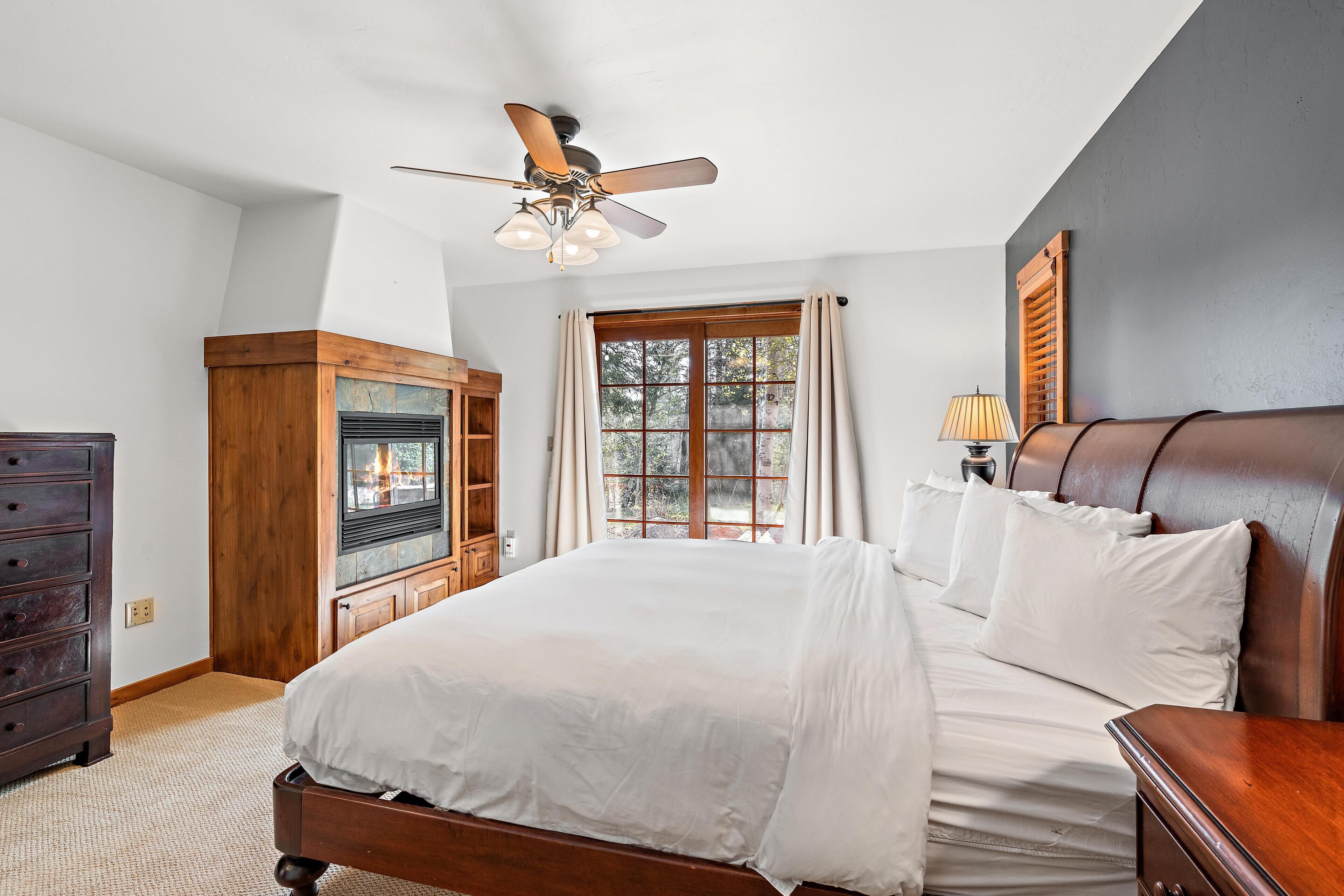 A bedroom with a large bed, a ceiling fan, a window with curtains, a wooden dresser, and a nightstand.