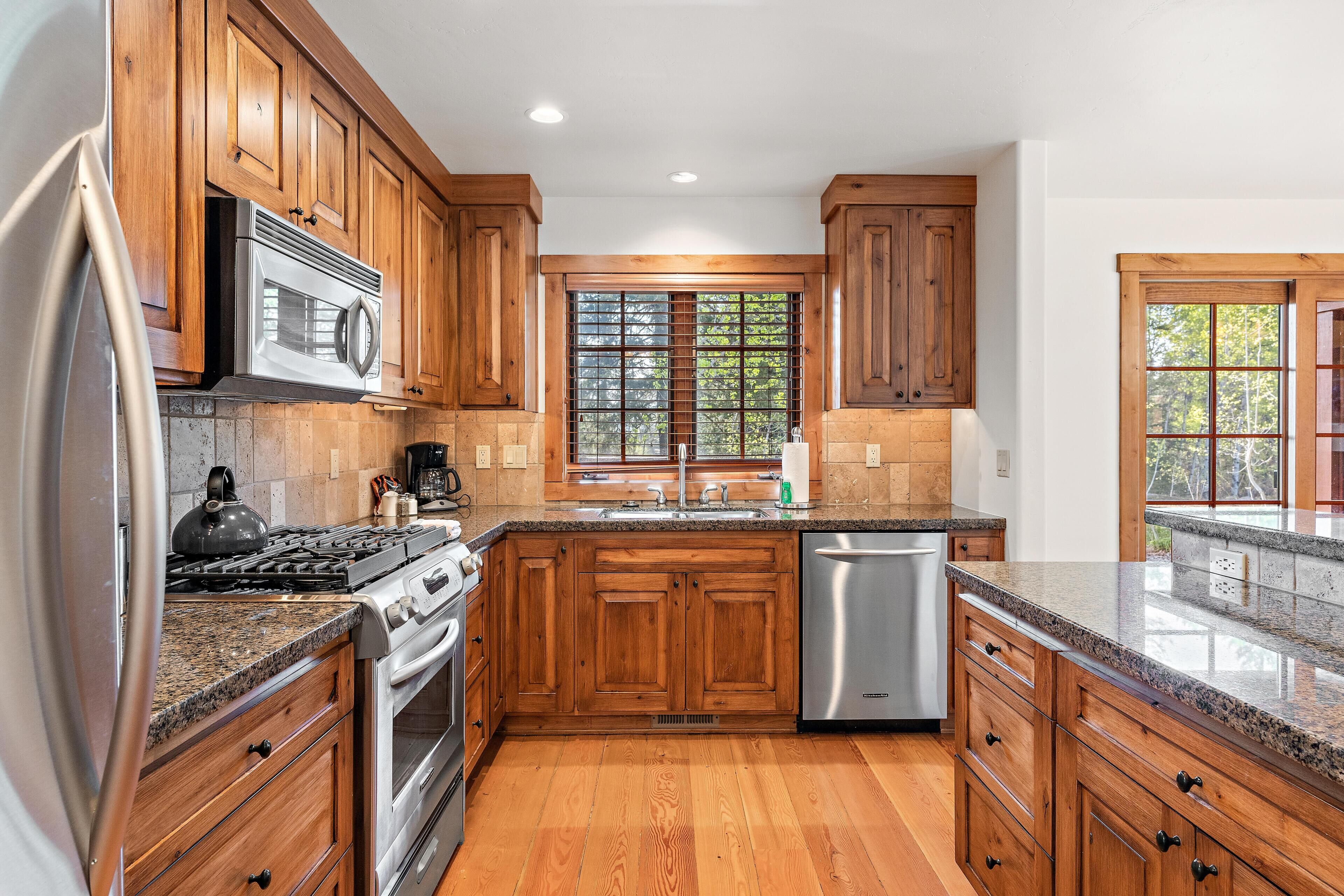 A kitchen with wooden cabinets, stainless steel appliances, a granite countertop, and a window with a view of trees.