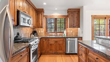 A kitchen with wooden cabinets, stainless steel appliances, a granite countertop, and a window with a view of trees.