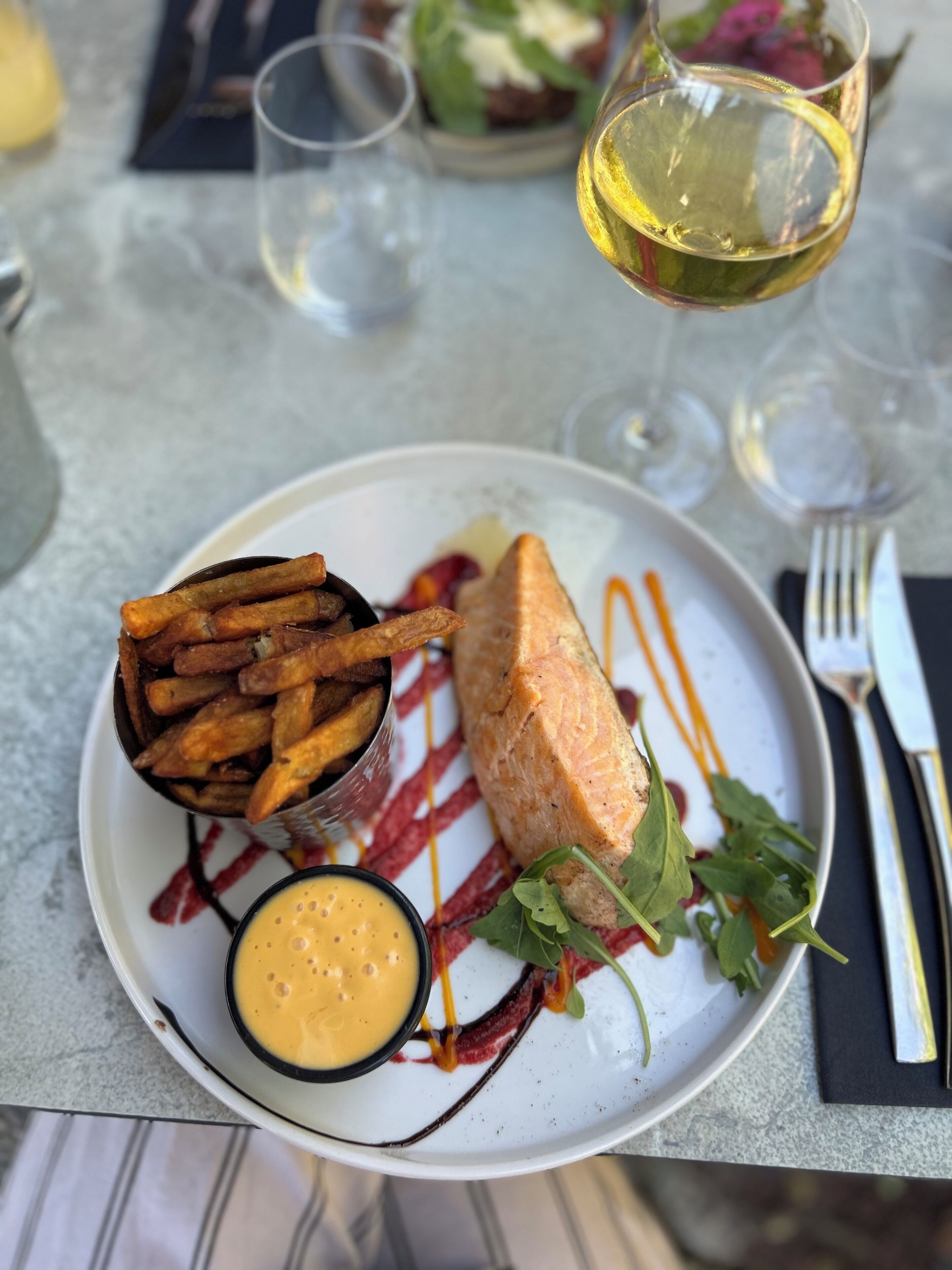 Une assiette contenant du saumon, des frites et une sauce, accompagnée d’un verre de vin et d’une fourchette, le tout posé sur une table.