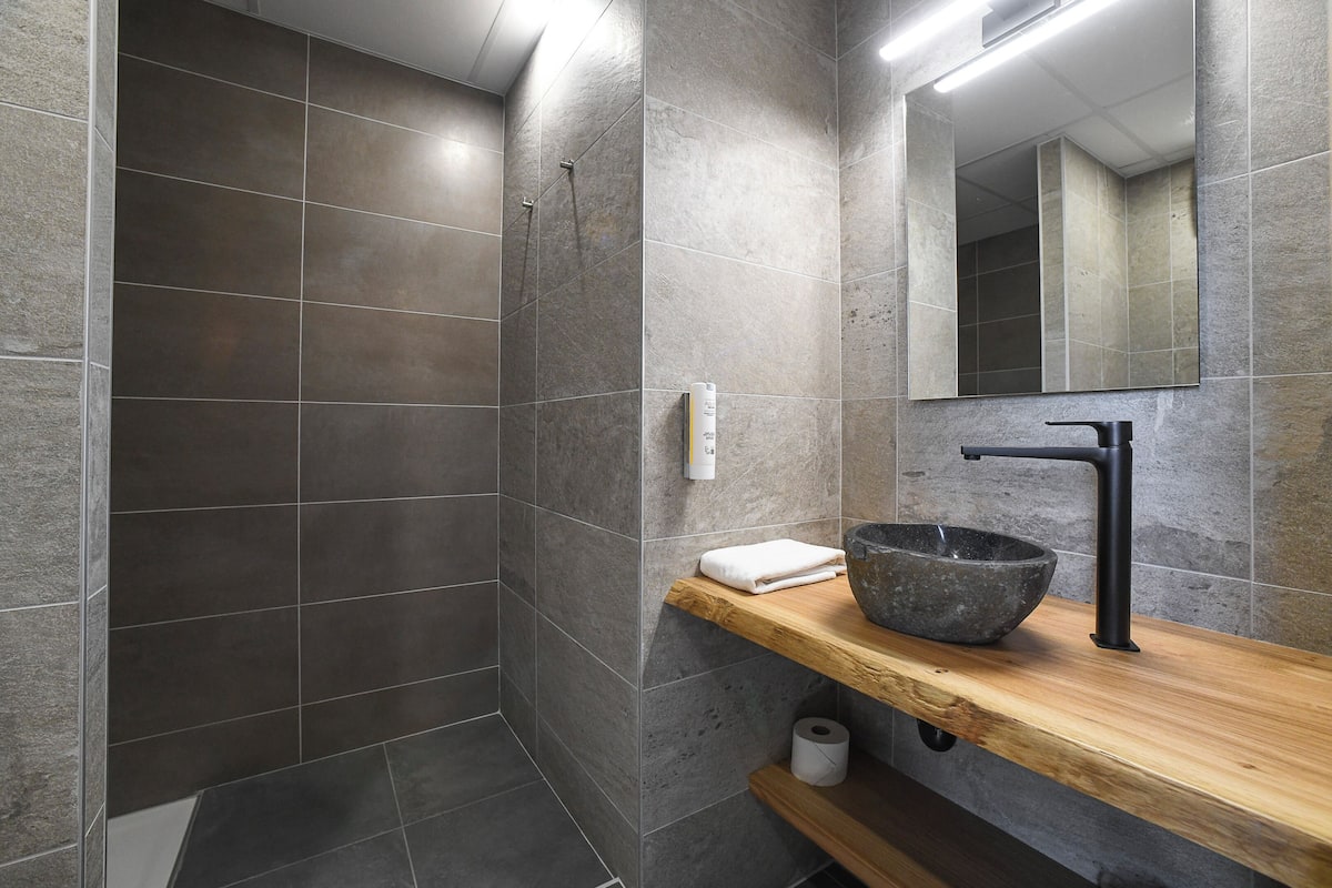 A modern bathroom with grey tiles, a wooden vanity, a black sink, and a mirror.