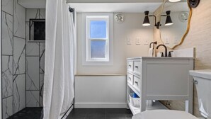Un baño con tocador blanco, inodoro, ducha con cortina blanca, ventana y espejo con luces integrado en la pared.