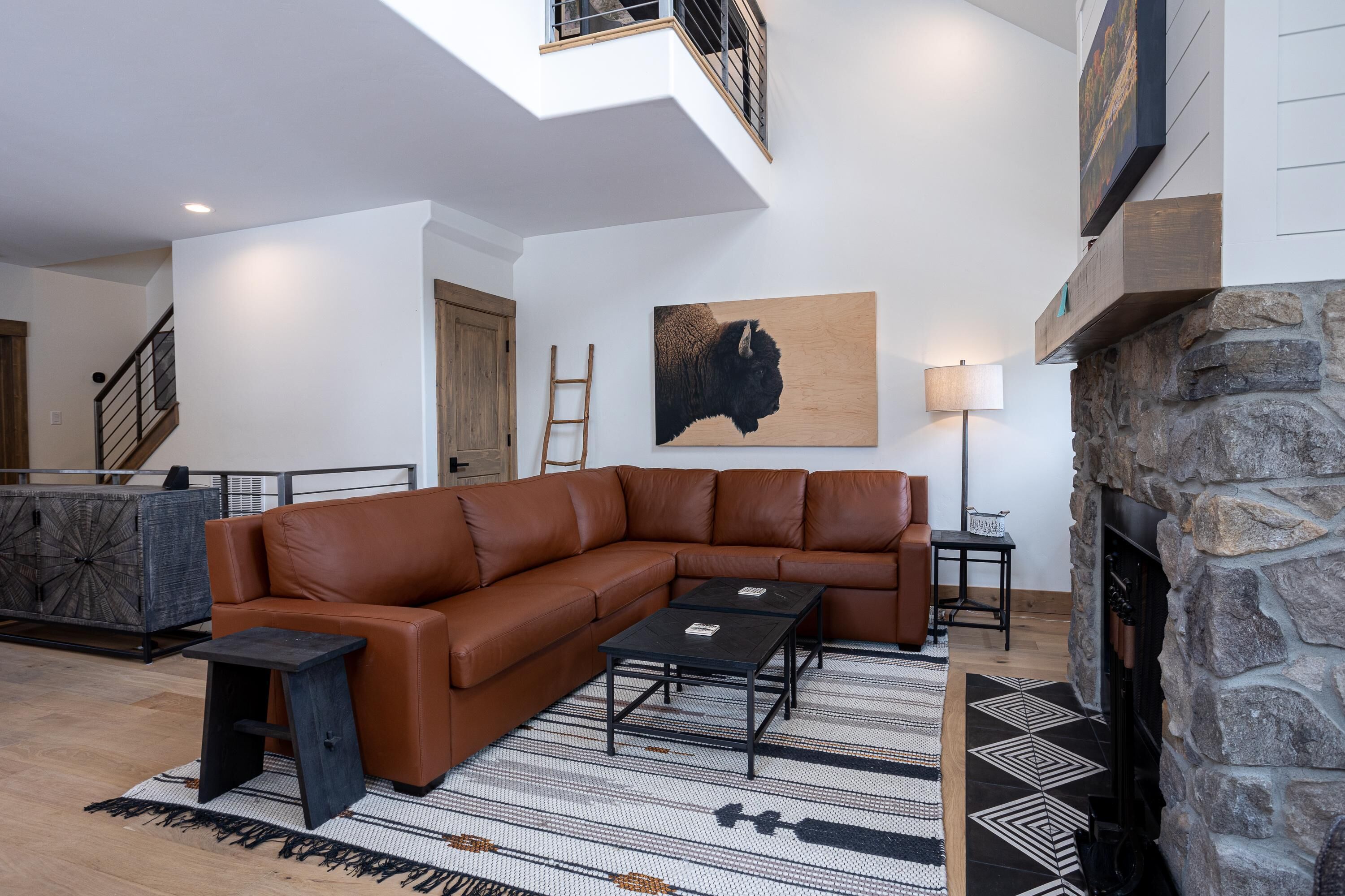 Un soggiorno moderno con un divano angolare in pelle marrone, due tavolini neri, un tappeto decorato, un caminetto in pietra e un grande quadro astratto appeso al muro.