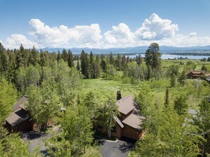 A cabin with a large deck, surrounded by dense forest and a view of mountains and a lake.