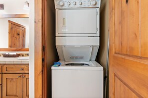 Une petite buanderie avec des lave-linge et des sèche-linge superposés, des armoires en bois et un plan de travail agrémenté d’un panier.