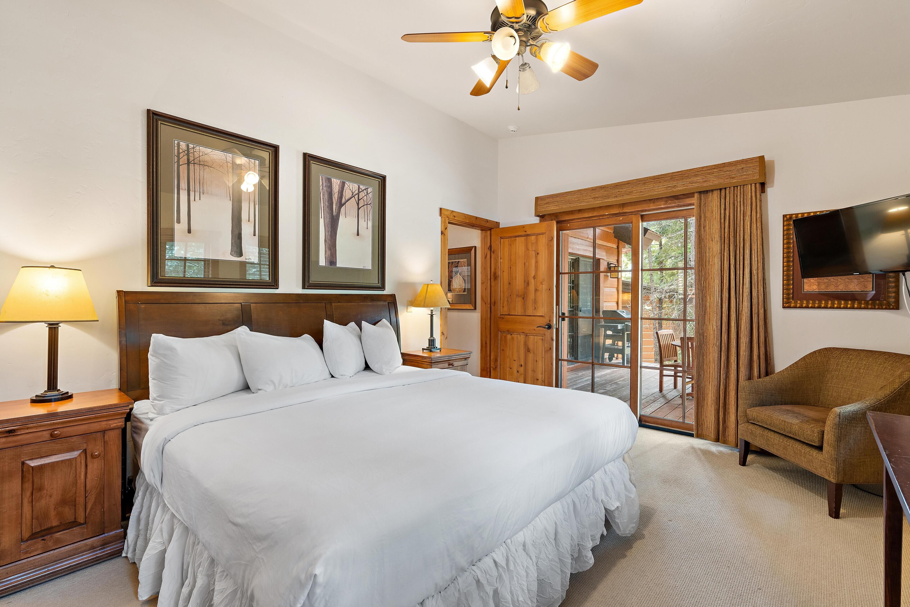 A hotel room with a large bed, wooden headboard, two bedside tables with lamps, a ceiling fan, a TV mounted on the wall, and a sliding glass door leading to an outdoor area.