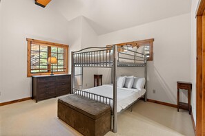 Een slaapkamer met een stapelbed, een dressoir, een raam met jaloezieën en een lamp.