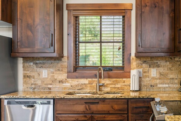 A kitchen with a granite countertop, wooden cabinets, a window with blinds, and a stainless steel dishwasher.